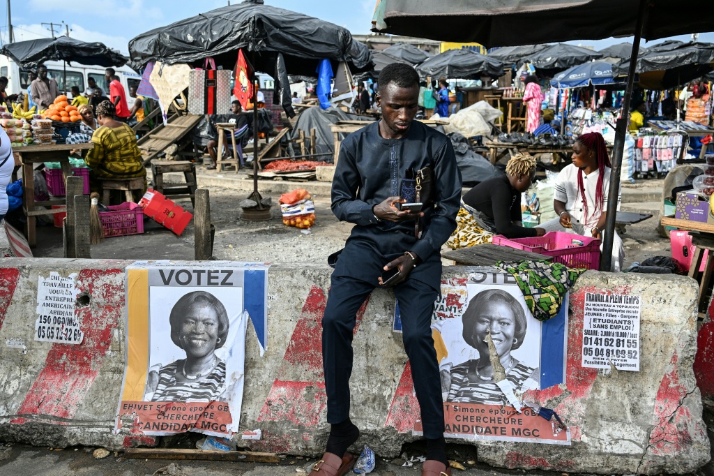 Un jeune homme est assis et regarde son téléphone, sur un bloc de béton sur lequel sont collées des affiches de campagne de l'opposante Simone Ehivet Gbagbo, à Yopougon, le 12 octobre 2025.